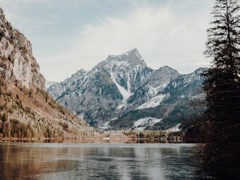 Ein teils zugefrorener See vor einem markanten, schroffen Berggipfel umringt von winterlich braunem Gras.