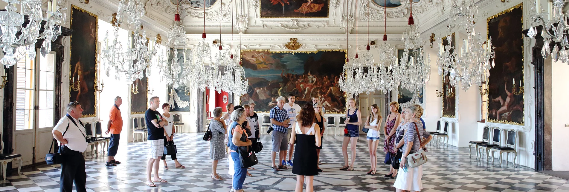 Gruppe von Besucherinnen und Besuchern bei einer Führung im prunkvollen Planetensaal von Schloss Eggenberg in Graz, mit barockem Deckenschmuck, Kronleuchtern und schwarz-weiß kariertem Marmorboden. | © Universalmuseum Joanneum