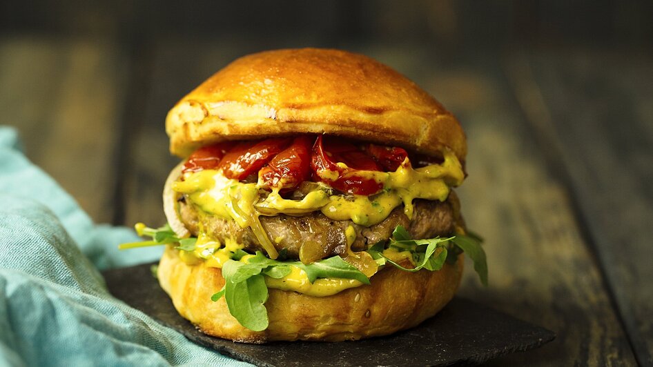 Saftiger Burger mit Salat, Tomaten und Saucen auf Holzbrett. | © TG-Graphics
