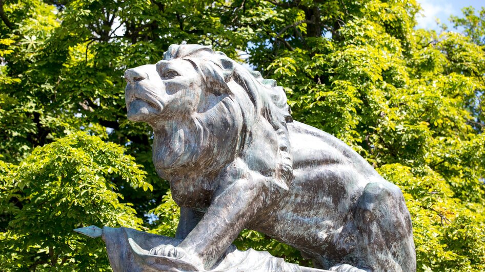 Bronze-Löwenskulptur auf dem Grazer Schlossberg vor grünen Bäumen.