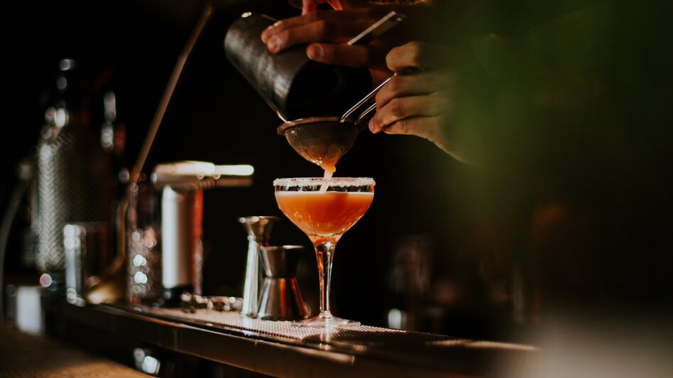 Ein Barkeeper gießt einen Cocktail in ein Glas an einer Bar. | © Tinksi