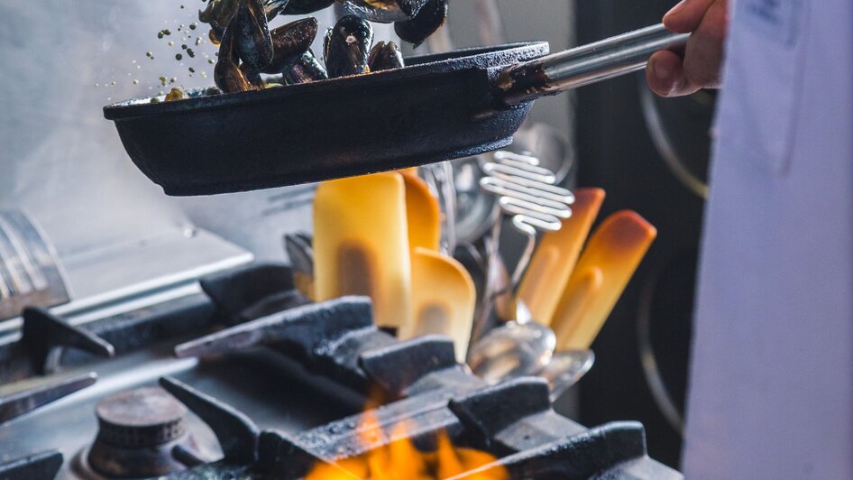 Koch hält eine Pfanne mit Muscheln über einen Ofen mit Feuer in einer Küche.
