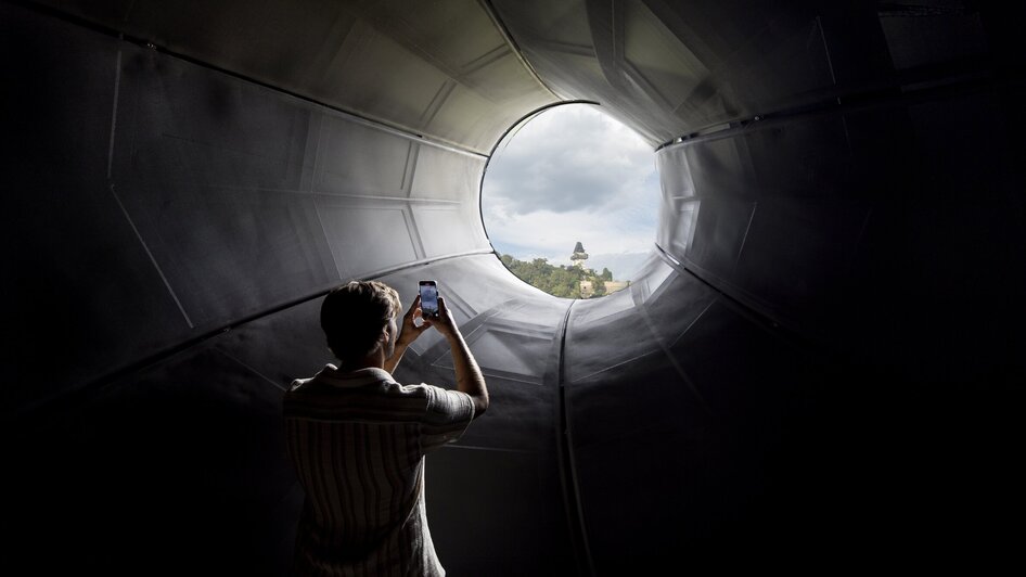 Besucher fotografiert den Grazer Uhrturm durch das Kunsthaus Graz.