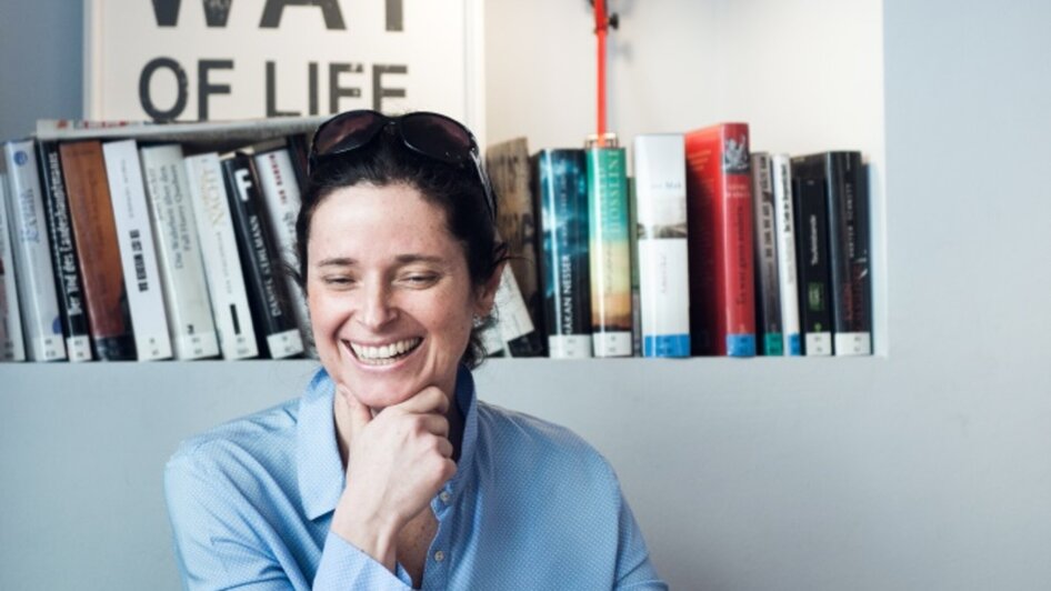 Lächelnde Frau in einem Café mit Bücherregal im Hintergrund.