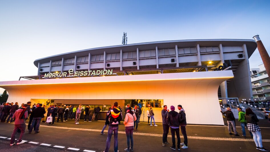 Besucher stehen vor dem Merkur Eisstadion in Graz bei Sonnenuntergang.