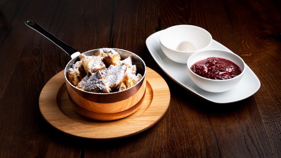 Kaiserschmarrn mit Staubzucker in einer kleinen Pfanne, daneben steht eine Schale mit Zwetschgenröster und eine leer Schale.