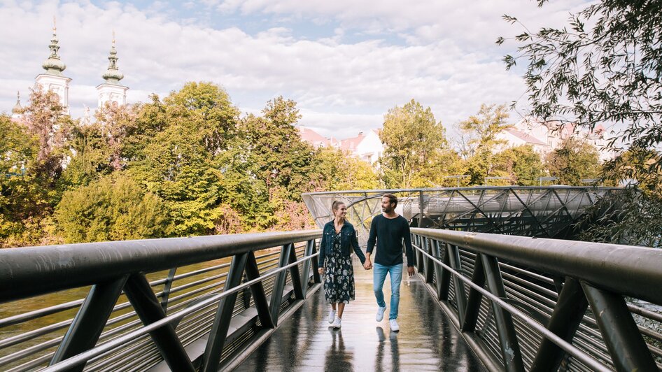 Paar spaziert auf der Murinsel in Graz, umgeben von Bäumen.