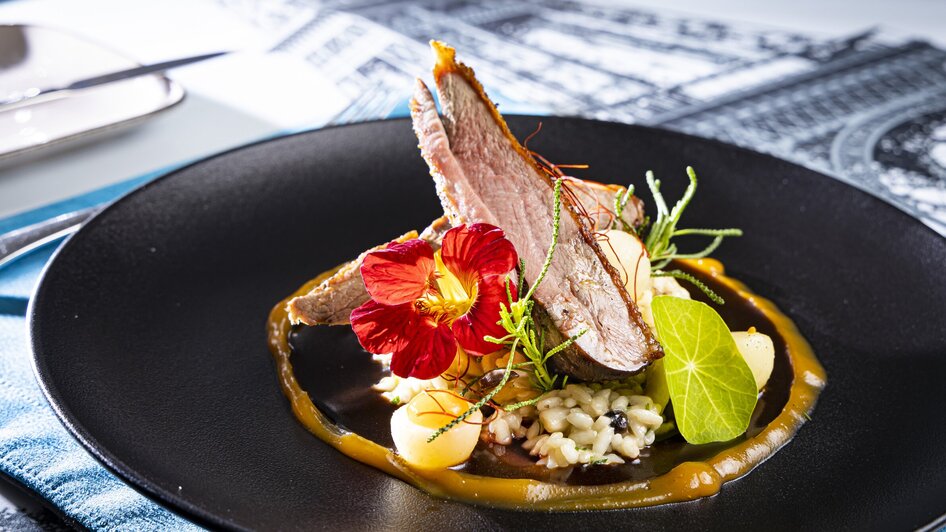 Ein kunstvolles Gericht mit Fleisch, Blüten und Risotto auf einem schwarzen Teller.