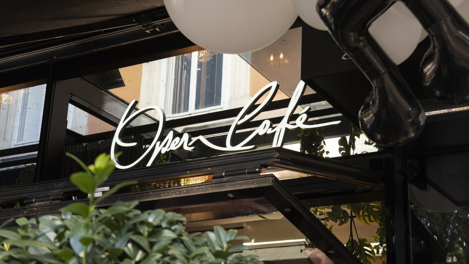 Beleuchtetes Schild des Operncafés mit Pflanzen im Vordergrund.