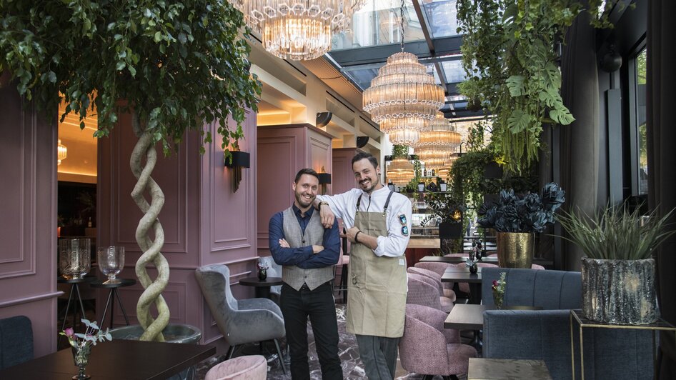 Zwei Männer im Operncafé, elegant dekorierter Raum mit Pflanzen und Lustern.