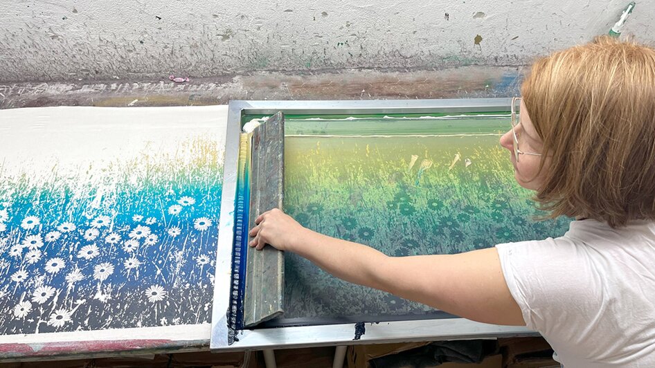 Eine Person arbeitet an einem Farbdruck im Atelier, umgeben von Kunstmaterialien. | © Peaces