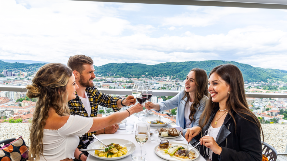 Im Schlossberg Restaurant sitzen vier Personen um einen Tisch mit Blick auf Graz. Sie stoßen mit Weingläsern an und genießen ein stilvolles Essen in einer modernen Umgebung. | © Paul Stajan