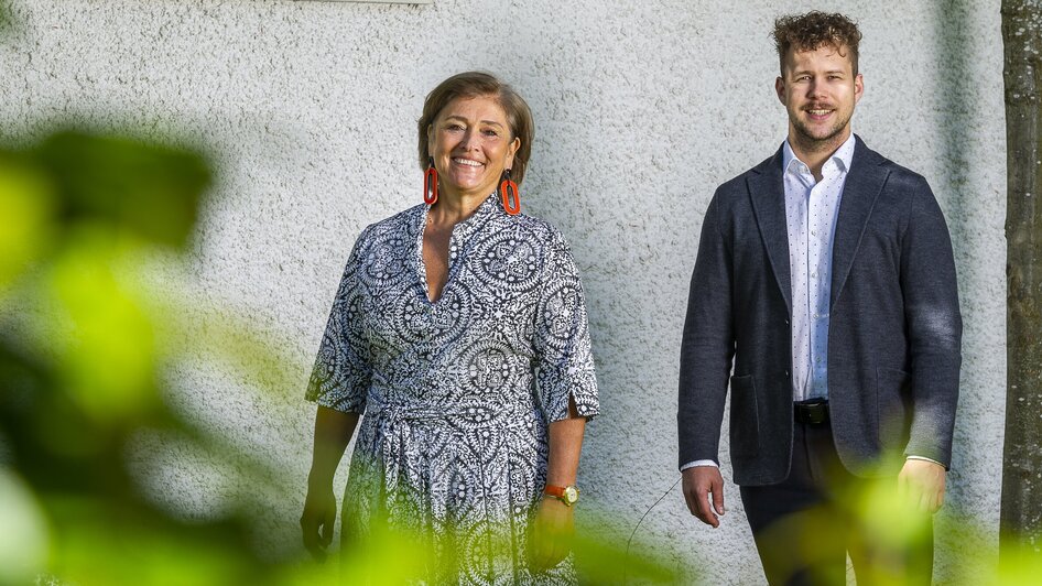Zwei Personen stehen in einem Garten, lächeln und posieren vor einer Wand.