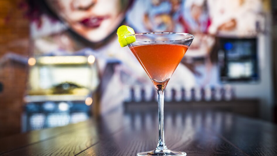 Ein eleganter Cocktail in einem Glas, dekoriert mit einer Limettenscheibe, vor einem Wandbild.