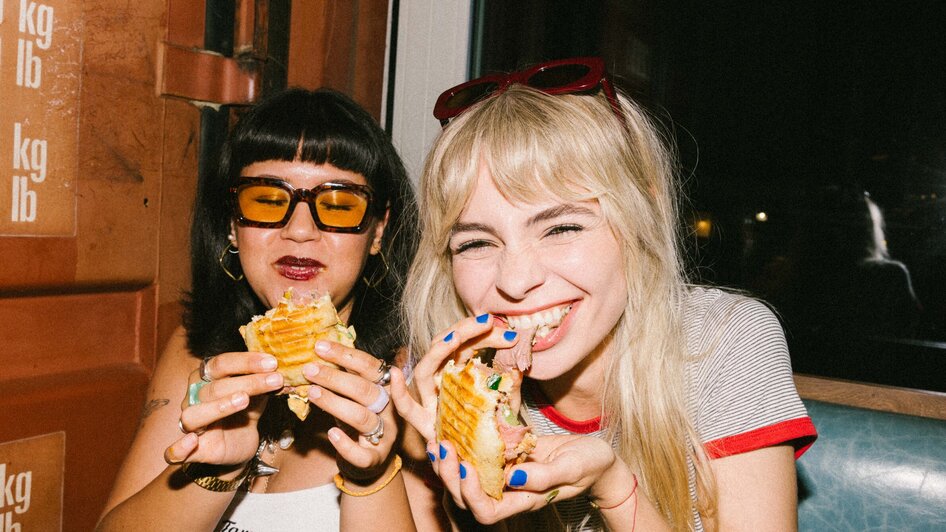Zwei Frauen genießen Sandwiches in einem Café und lachen. | © Bobby's Agency - Simon Bauer