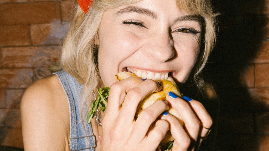 Eine Frau lächelt und genießt einen Burger, während sie breit grinst. | © Bobby's Agency - Simon Bauer