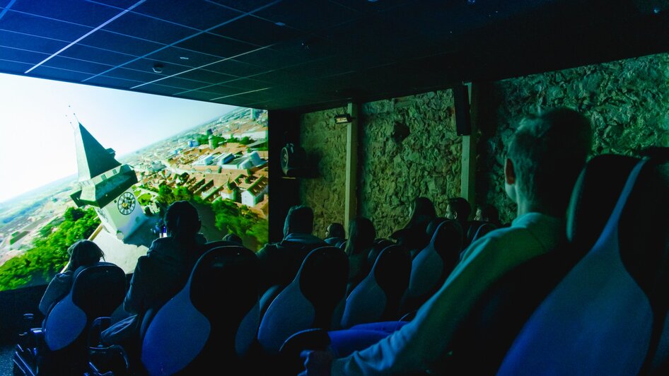Besucher im 4D Kino im Grazer Schlossberg mit Grazer Uhrturm auf der Kino-Leinwand.