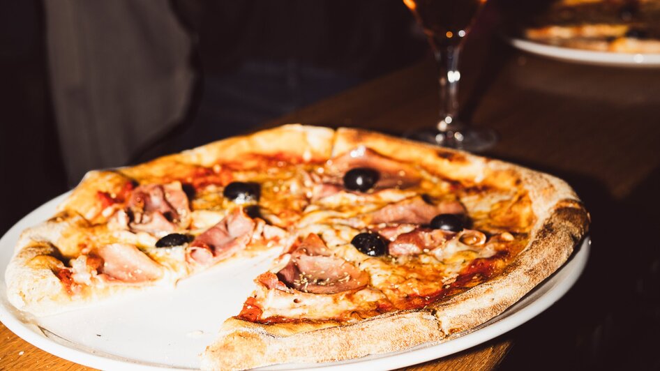 Ein Teller Pizza mit Oliven und Schinken, dazu ein Drink auf einem Tisch in einer Bar.