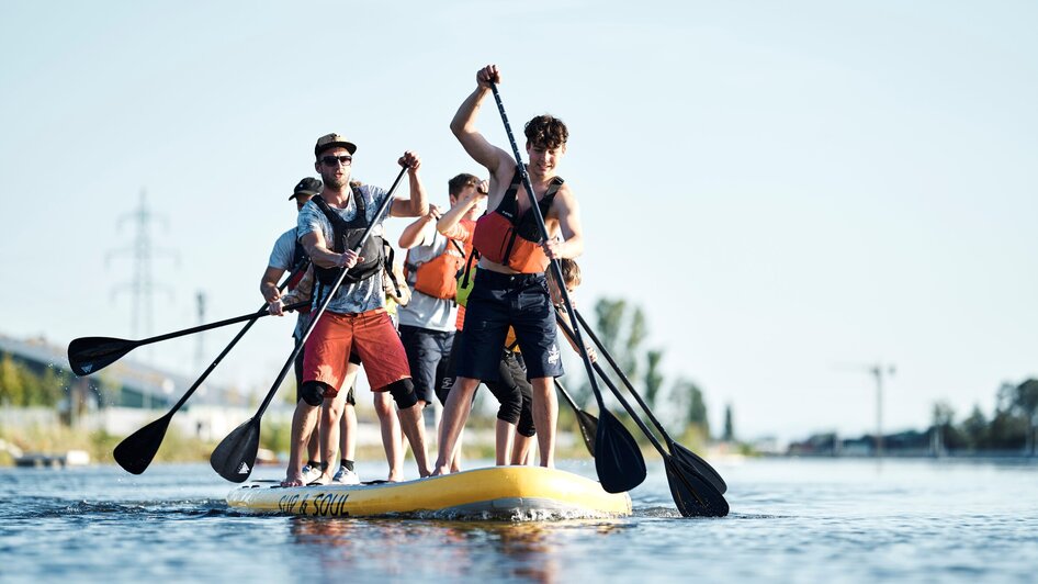 Boot-Verleih am Stadtstrand in Graz | © Stadtstrand Graz - Stefan Leitner Gruppe von Menschen paddelt auf einem großen SUP im Wasser. | © Stadtstrand Graz - Stefan Leitner