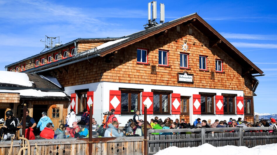 Winterlich verschneite Berghütte mit Holzfassade und zahlreiche Menschen genießen bei Sonnenschein regionale Speisen und Getränke auf der Terrasse | © Archiv Hochwurzenhütte