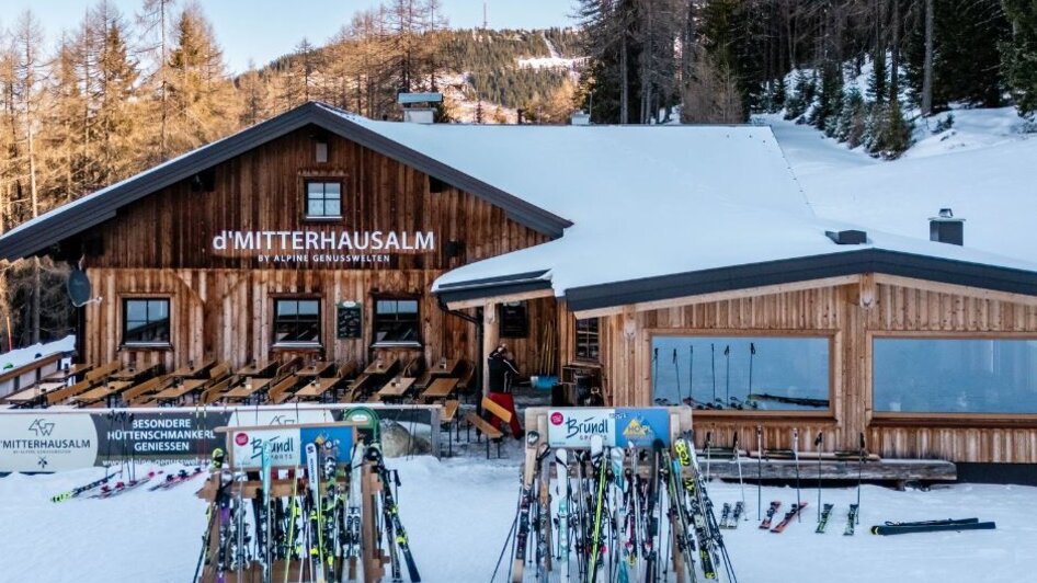 Holzhütte im Schnee mit Terrasse und vielen abgestellten Skiern davor, umgeben von Bäumen. | © Mitterhausalm | Alpine Genusswelten