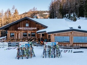 Holzhütte im Schnee mit Terrasse und vielen abgestellten Skiern davor, umgeben von Bäumen. | © Mitterhausalm | Alpine Genusswelten