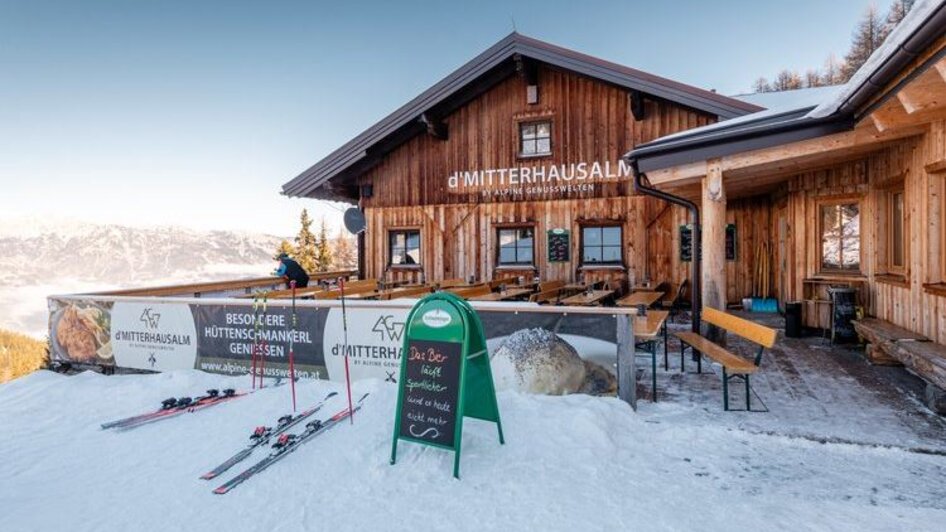 Holzhütte im Schnee mit Terrasse, Skiern davor und Bergblick im Hintergrund. | © Mitterhausalm | Alpine Genusswelten